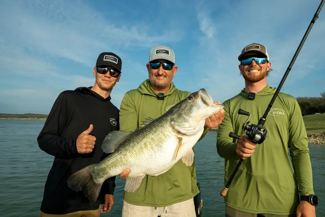 Three U-Call anglers in U-Call hooded shirts and caps holding a largemouth bass